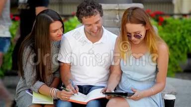 一个英俊的男孩和两个漂亮的女孩在一起聊天和学习户外坐在长凳上温暖的阳光